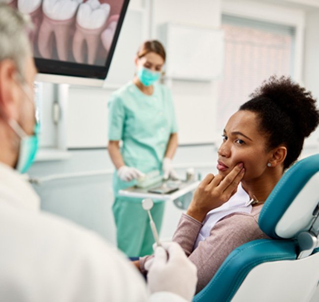 Woman with brown curly hair touching two fingers to her jaw in pain sitting in teal dental chair