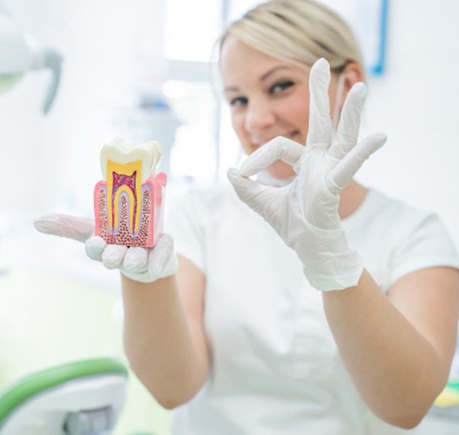 Dentist in white gloves and scrubs holding up model tooth making “OK” sign with fingers